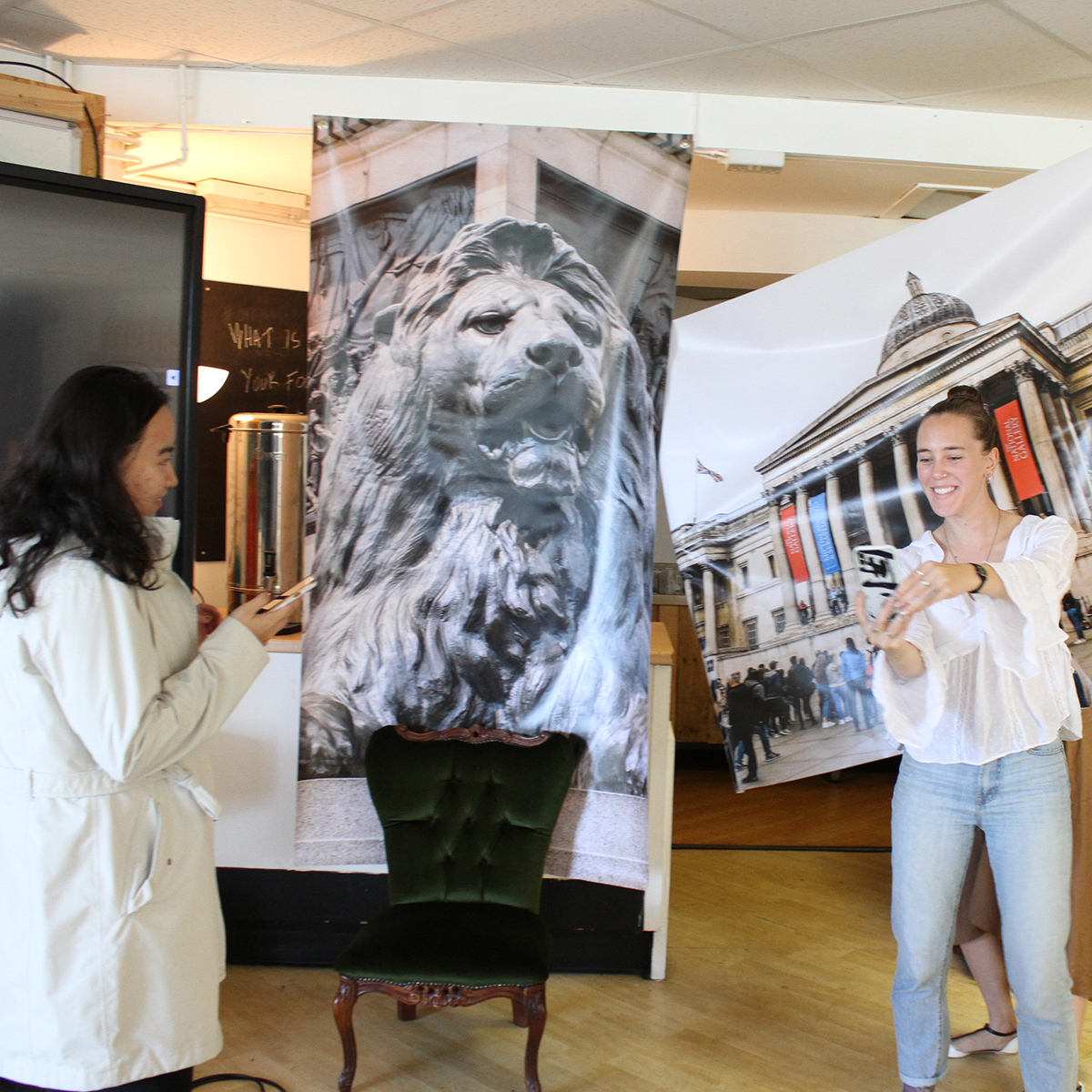 Two students taking selfies in front of large posters of london scenes