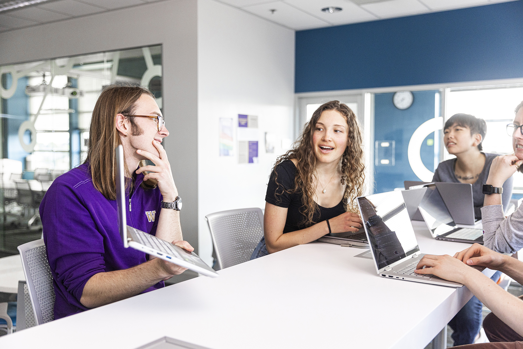 Five students sit around a table on laptops. One student in a UW shirt holds up their laptop screen toward the group.