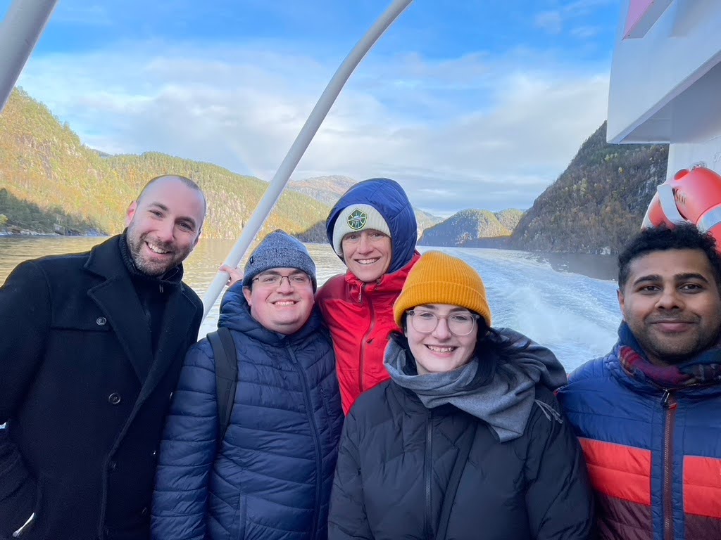 A group stands on a ferry boat smiling, Fjords are visible in the background