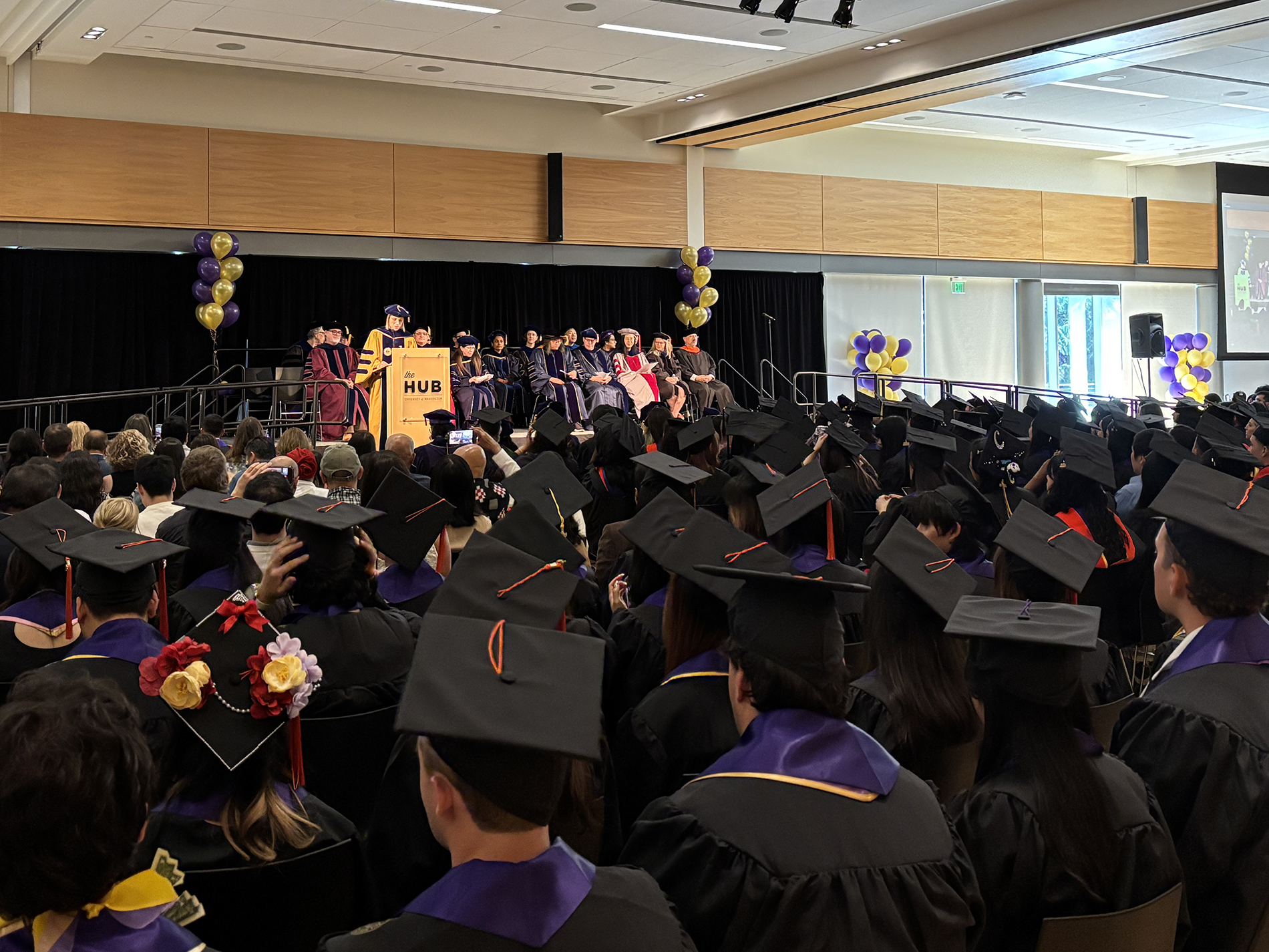 Julie Kientz addresses the room from the podium. Graduates are seated wearing regalia in the foreground.
