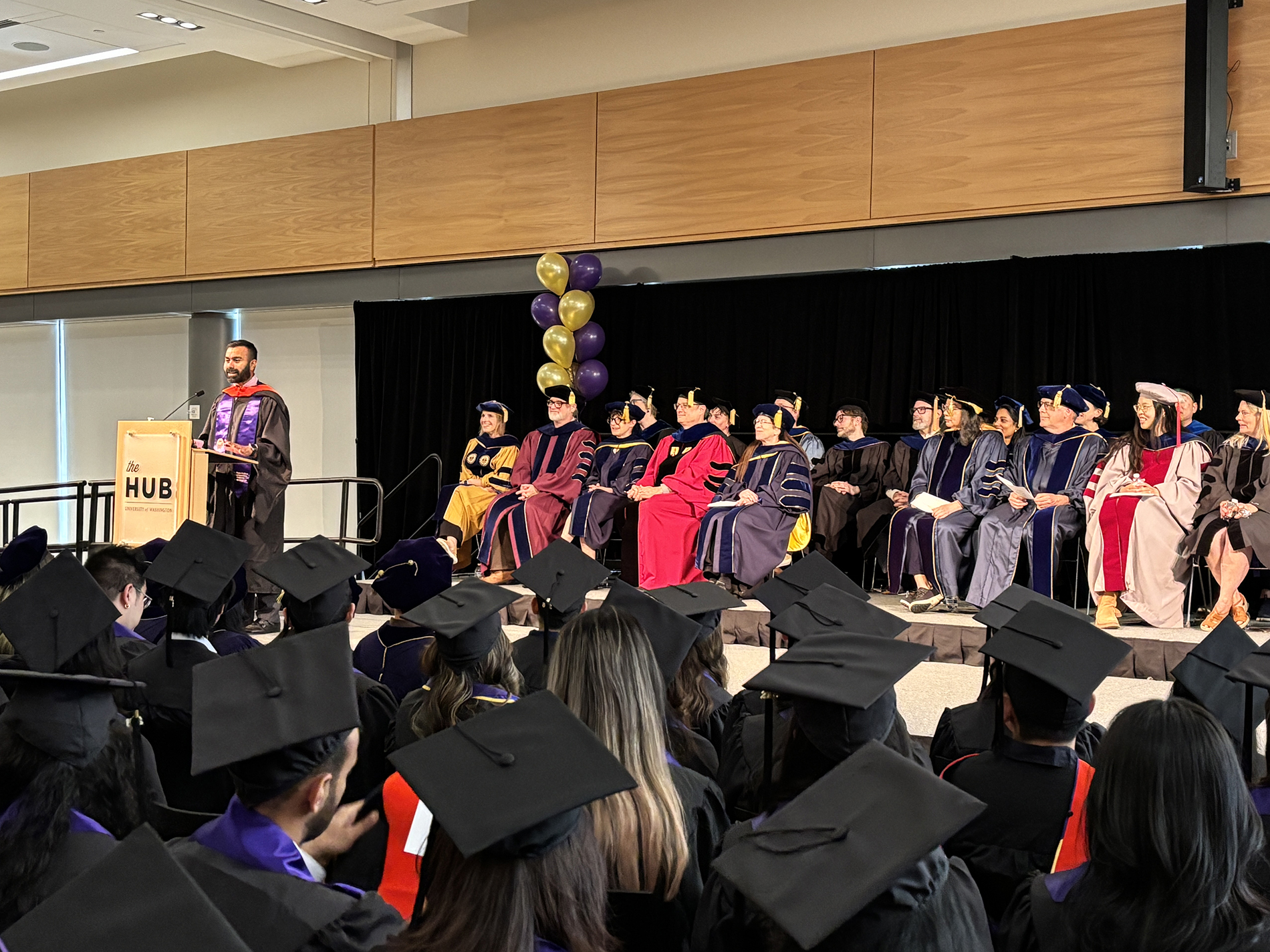Ram Shankar Siva Kumar wears graduation regalia and stands at the podium in front of a room full of graduates with HCDE faculty seated behind him