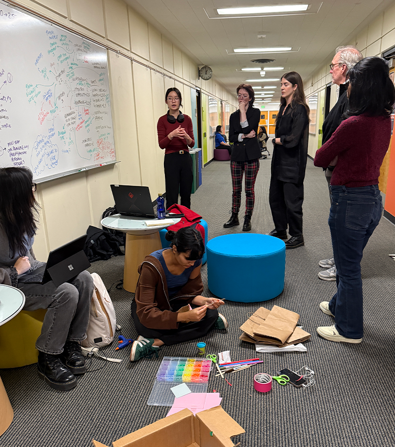 Students and mentors gather in the hallway of Sieg Building around a whiteboard. Prototyping supplies like cardboard and sticky notes are on the floor in front of them.