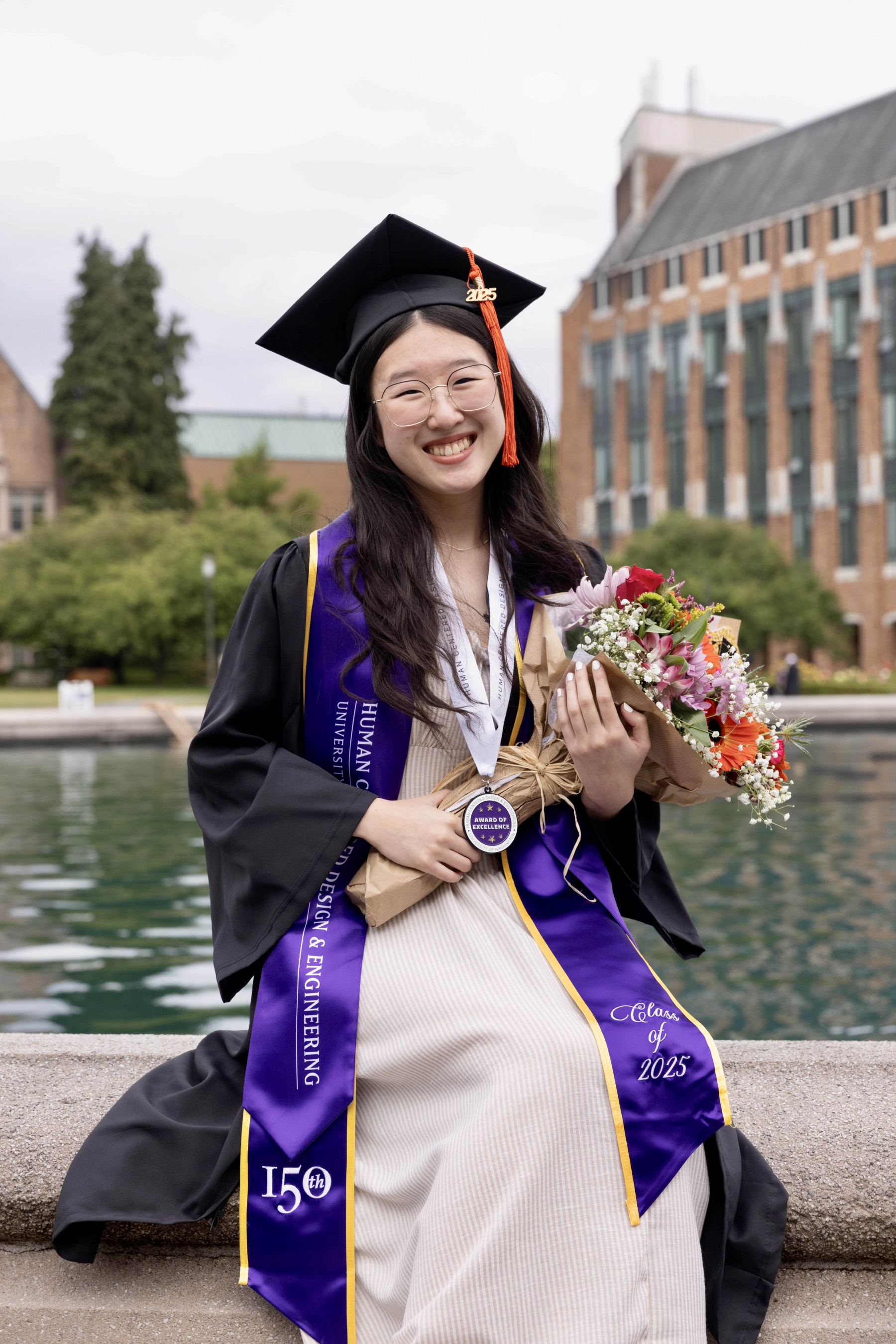 Camryn Soo wears her UW graduation regalia, sitting along the edge of the UW fountain and holding flowers