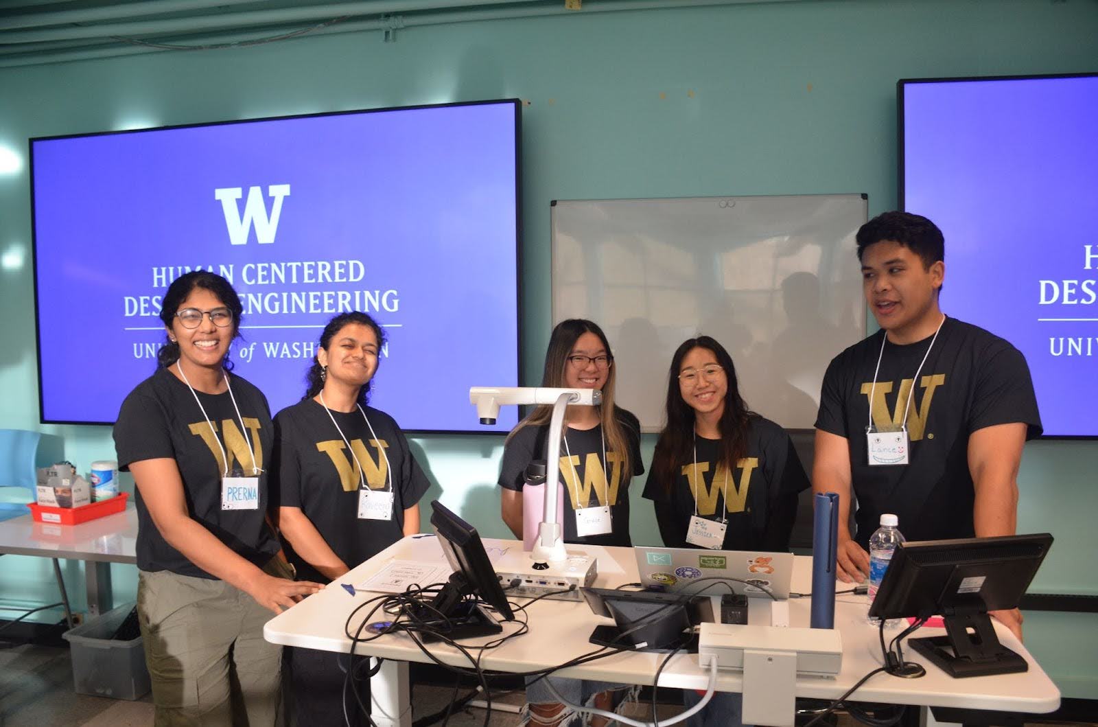 Five UW students in HCDE t-shirts stand in front of a classroom in front of HCDE logos on screen
