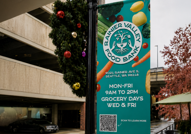 Rainier Valley Food Bank street banner concept featuring the food bank logo, grocery hours, address and a QR code, shown hanging from a neighborhood light pole.