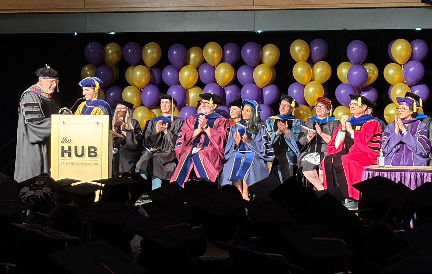 James Landay receives the award from Julie Kientz on stage at the 2025 HCDE Graduation Ceremony with HCDE faculty seated and clapping behind them.