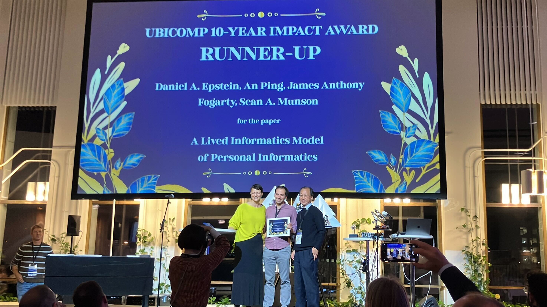 Daniel Epstein on stage at UBICOMP holds an award with a slide saying: UBICOMP 10-YEAR IMPACT AWARD RUNNER UP presented on screen behind him