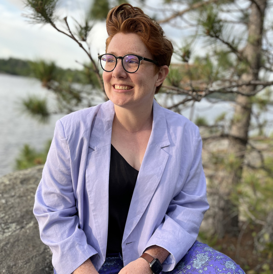 Emma McDonnell, a white woman with short red hair and circular black glasses sits on a rock with a lake blurred in the background behind her, wearing a lavender blazer, black tank top, and floral patterned purple pants. She is not looking directly at the camera, but is smiling looking out.