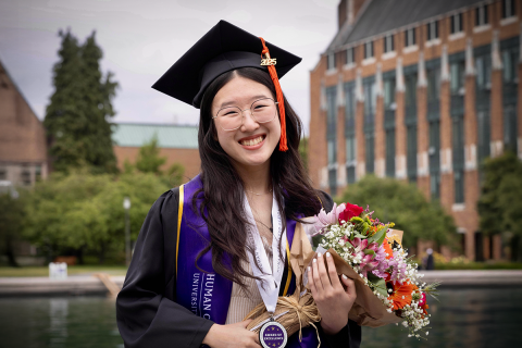 Camryn Soo wears her UW graduation regalia, sitting along the edge of the UW fountain and holding flowers