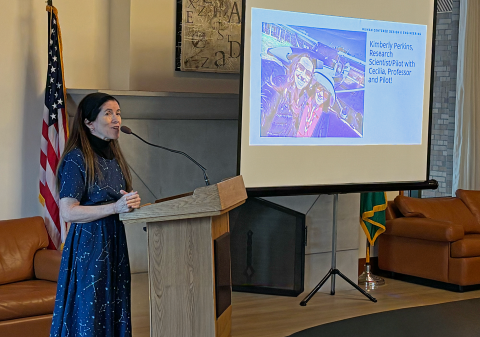 Cecilia Aragon stands behind a podium speaking to the room at her retirement party. A slideshow with a photo of Cecilia and a student is on the screen behind her.