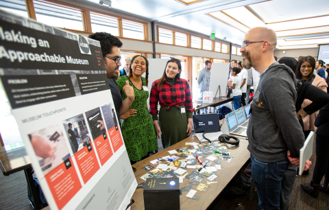 Three students stand with their project poster at the capstone showcase talking with a visitor