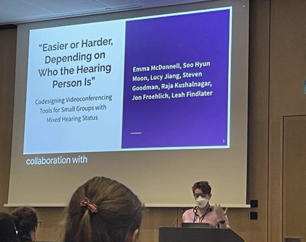 Emma, a white woman with red hair, presents her paper at CHI 2023. Behind her on screen is the title slide for her talk, with the paper’s title and collaborators listed.