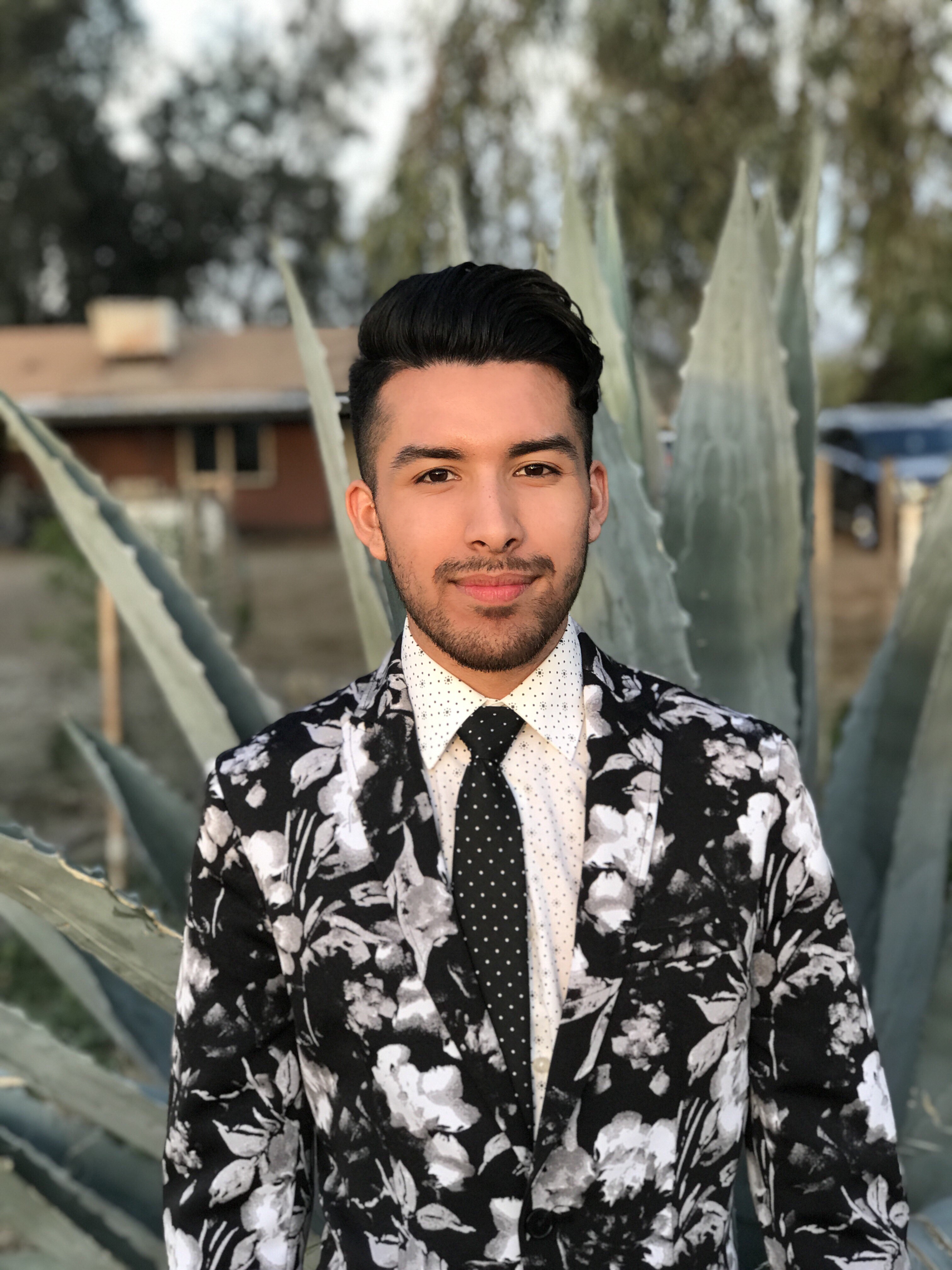 Alfie Aguilar Vidrio, standing in front of a cactus, wears a black and white floral-print suit jacket and smiles at the camera