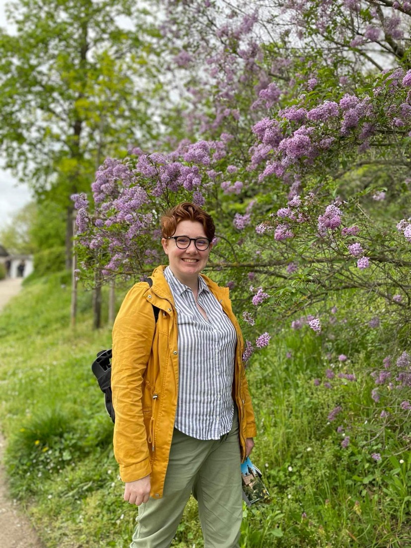 Emma, a white woman with short red hair wearing round black glasses, stands in front of a lilac bush, smiling at the camera. Her yellow coat and blue and white striped button down pop against the bushes behind her on an overcast day.
