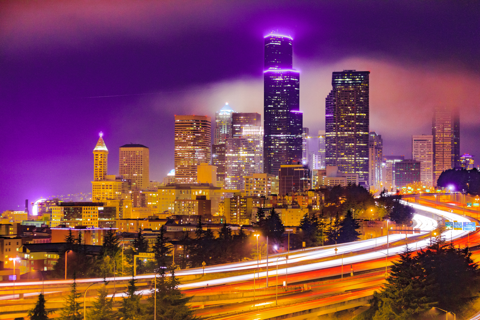 Seattle city scape at night with I-5 in the foreground. A purple sky is behind the buildings.
