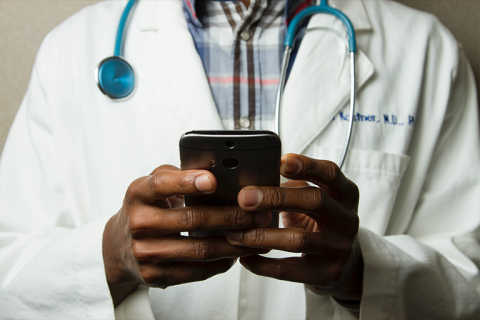 Close up of a doctor's hands holding a phone. The doctor is wearing a white coat and a stethoscope around their neck