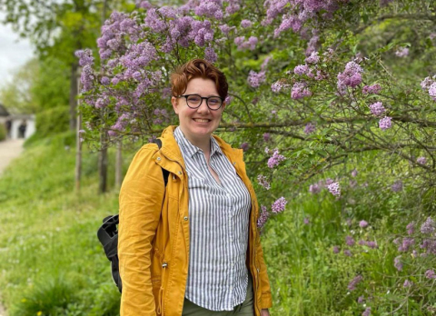 Emma, a white woman with short red hair wearing round black glasses, stands in front of a lilac bush, smiling at the camera. Her yellow coat and blue and white striped button down pop against the bushes behind her on an overcast day.